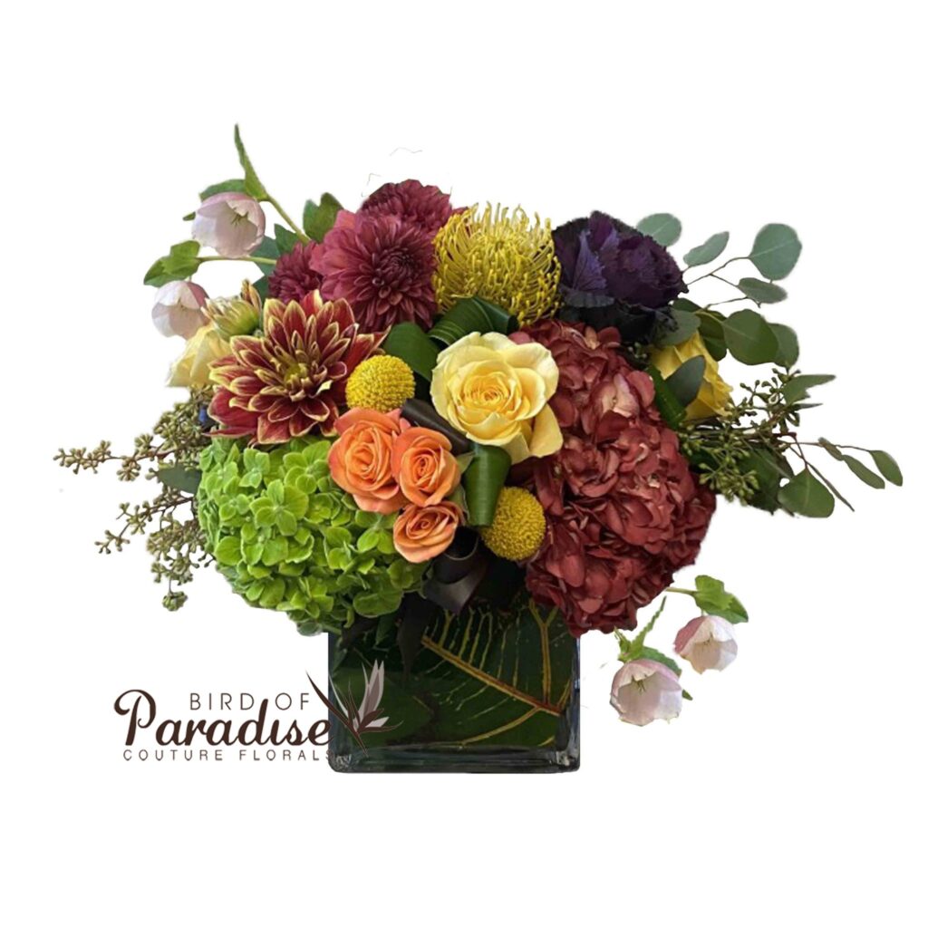 This flower arrangement will be the jewel of your fall table with dusky colors perfect for the season! This arrangement includes hydrangeas, roses, dahlias, cabbage, spray roses, pincushions, billy balls, and a variety of greenery.  All arranged in a square glass vase lined with foliage.