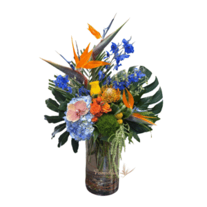 This blue tropical flower arrangement includes hydrangea, delphinium, birds of paradise, roses, spray roses, billy balls, dianthus, orchids, pincushions, and greenery arranged in a glass cyllinder vase.