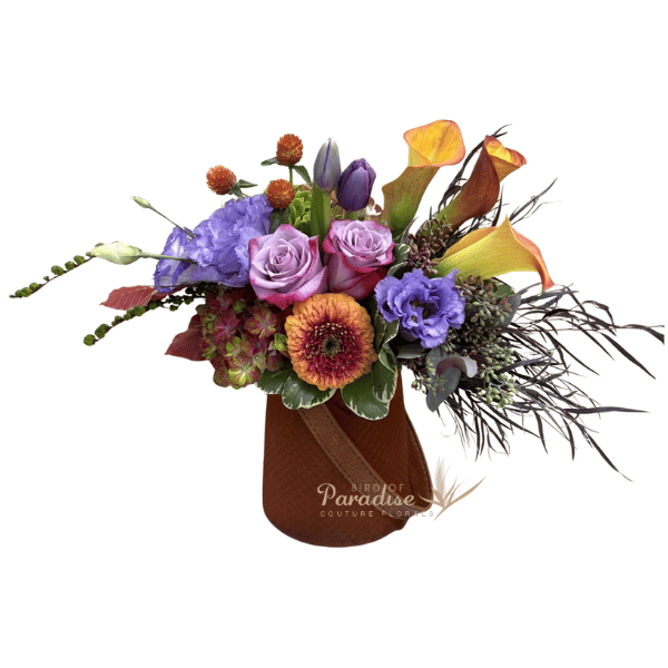 A myriad of purpe blooms usher in the fall seson in this purple fall flower arrangement.. It includes roses, gerbera daisies, hydrangeas, lisianthus, calla lilies, tulips, greenery and fall accents,  It's arranged in an orange glass vase with a decorative leather strap. 